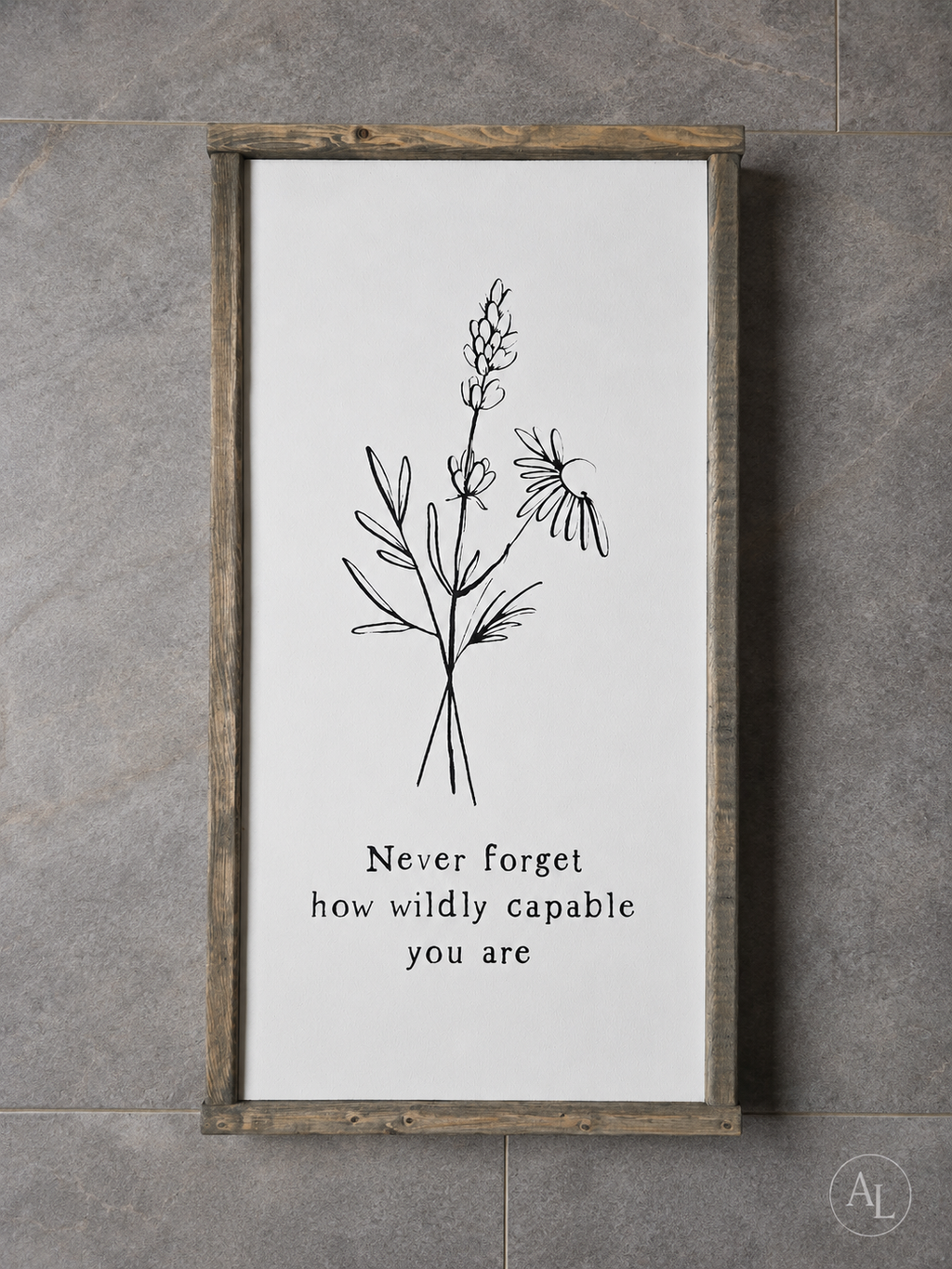A framed wall art print with a rustic wood frame, featuring a black line drawing of wildflowers and the text 'Never forget how wildly capable you are' on a white background, mounted against a gray tiled wall.