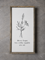 A framed wall art print with a rustic wood frame, featuring a black line drawing of wildflowers and the text 'Never forget how wildly capable you are' on a white background, mounted against a gray tiled wall.