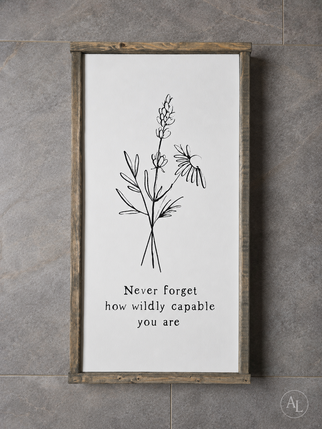 A framed wall art print with a rustic wood frame, featuring a black line drawing of wildflowers and the text 'Never forget how wildly capable you are' on a white background, mounted against a gray tiled wall.