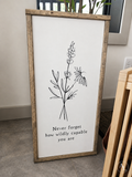 A framed vertical wall art print with a rustic light brown wood frame, featuring a black line drawing of wildflowers and the text 'Never forget how wildly capable you are' on a white background, leaning against a wall in an indoor setting.