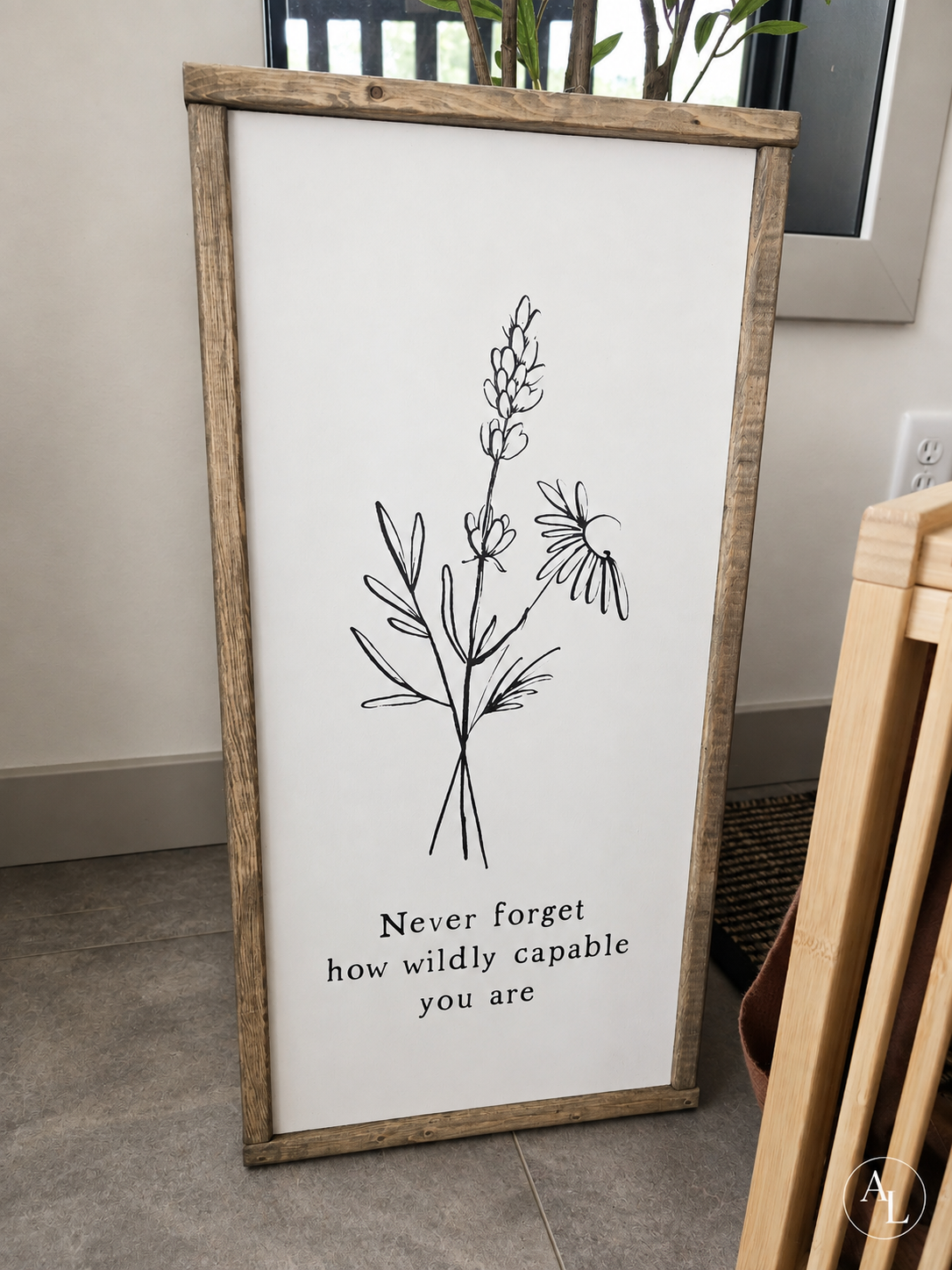 A framed vertical wall art print with a rustic light brown wood frame, featuring a black line drawing of wildflowers and the text 'Never forget how wildly capable you are' on a white background, leaning against a wall in an indoor setting.