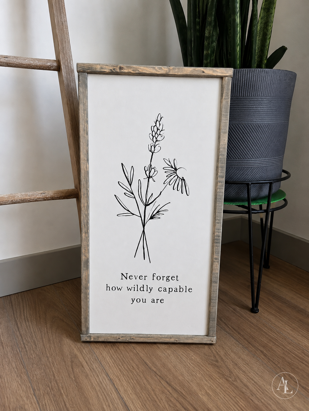 A framed vertical wall art print leaning on a wood floor, featuring a black botanical line drawing and the text 'Never forget how wildly capable you are' on a white background. A potted snake plant and a wooden ladder are visible in the room setting.