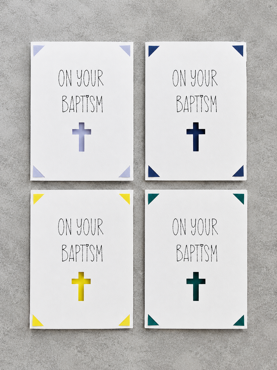 Four square wall art prints arranged in a 2x2 grid on a gray textured background. Each print has the words 'ON YOUR BAPTISM' in black lettering with a cross symbol below, and the corners are decorated with different colored triangle accents in light blue, navy, yellow, and teal.