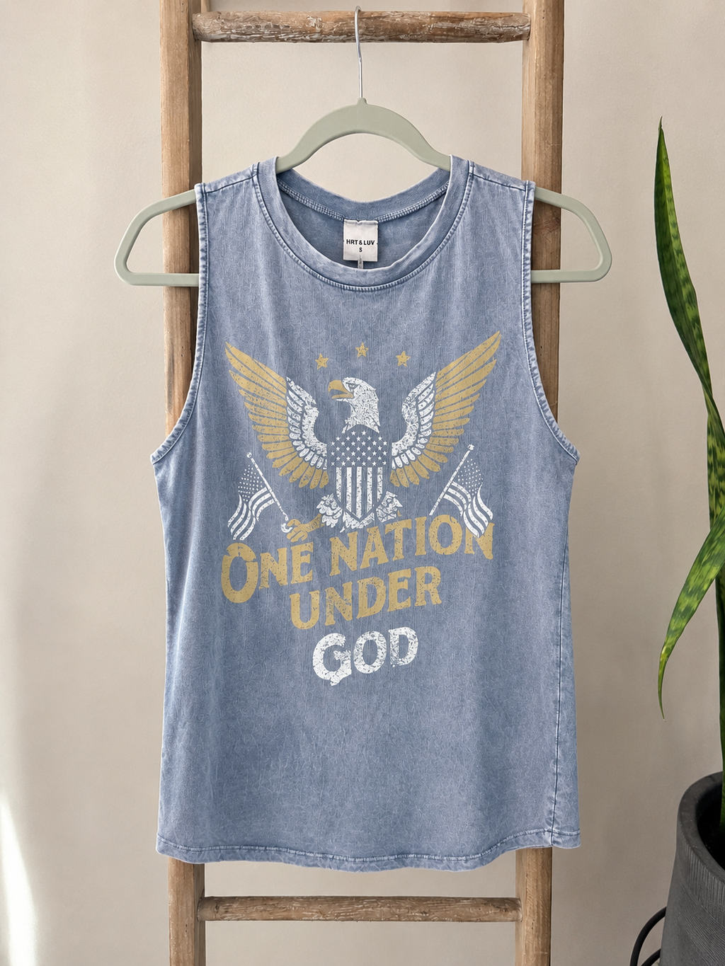 A light blue washed tank top hanging on a hanger against a beige wall, featuring a distressed graphic of an eagle with gold wings, American flags, and the text 'ONE NATION UNDER GOD'.