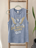 A light blue washed tank top hanging on a hanger against a beige wall, featuring a distressed graphic of an eagle with gold wings, American flags, and the text 'ONE NATION UNDER GOD'.