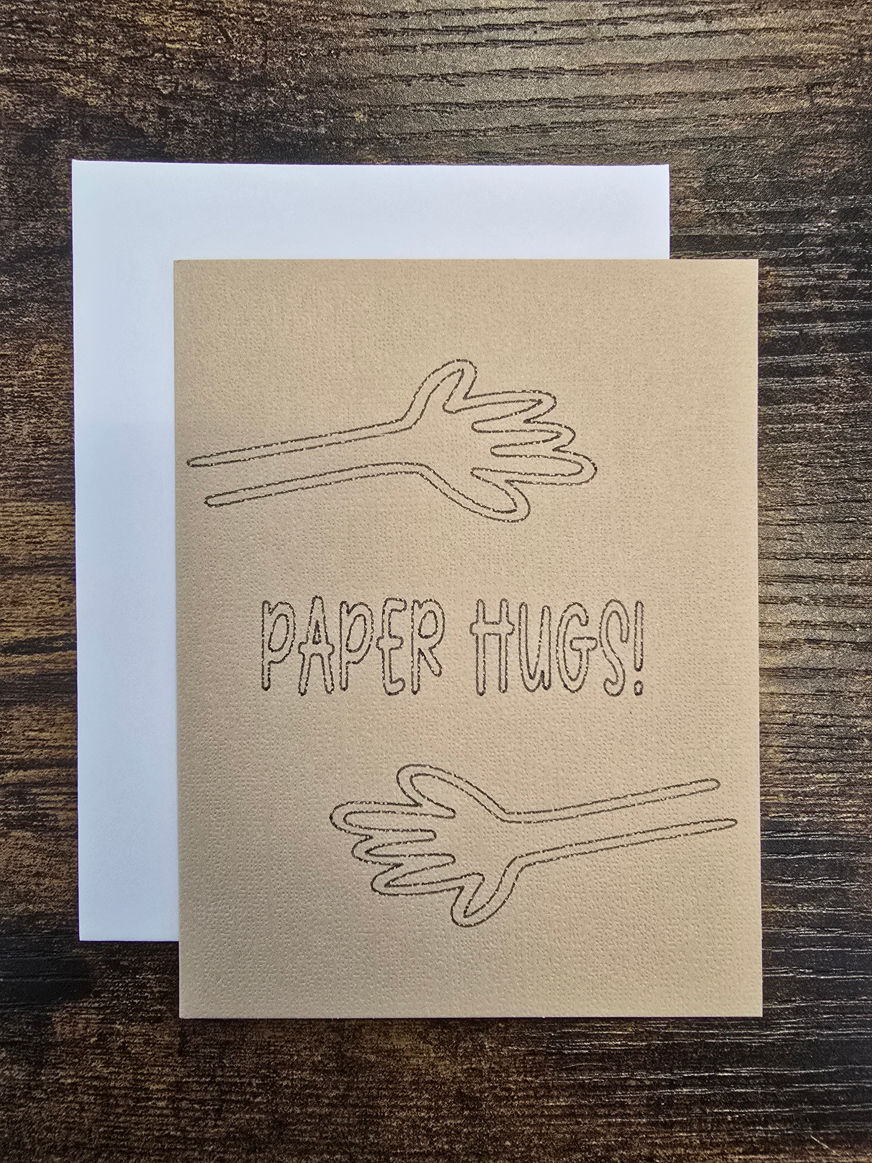 A square kraft paper greeting card with the words 'PAPER HUGS!' and two simple line drawings of hands, shown with a white envelope on a dark wood surface.