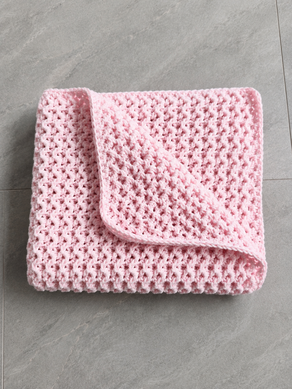 A folded light pink crocheted blanket with a textured openwork pattern, displayed on a gray tiled floor.