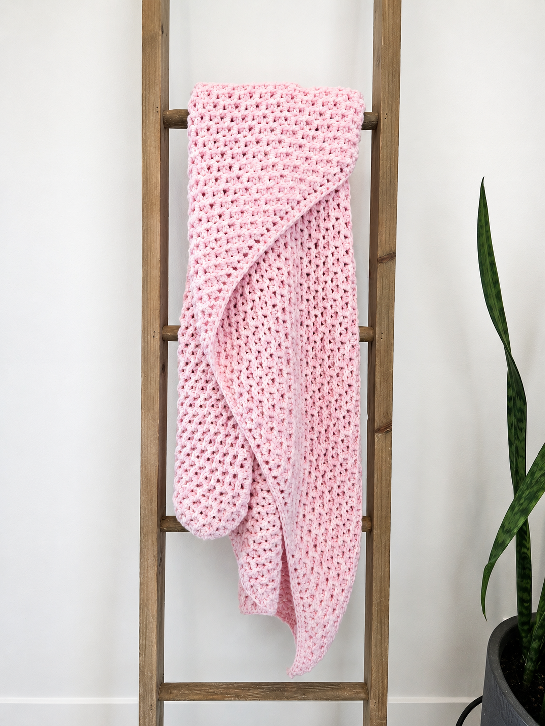 A light pink crocheted blanket with a hood is draped over a rustic wooden ladder against a white wall, with a green potted plant visible on the right.