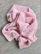 A light pink crocheted blanket with a textured open-knit pattern, casually draped on a gray tiled floor.