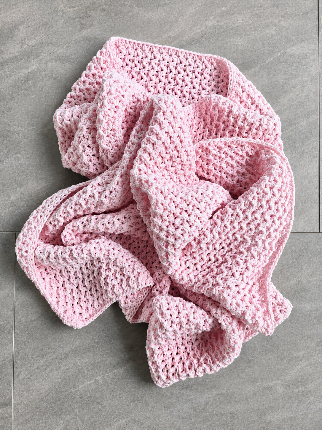 A light pink crocheted blanket with a textured open-knit pattern, casually draped on a gray tiled floor.