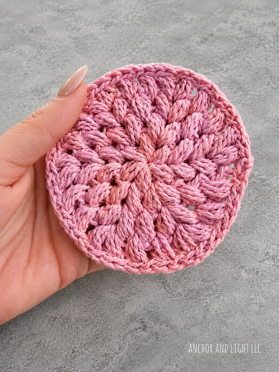 A hand holds a round crocheted coaster in a dusty rose pink color against a textured gray background.