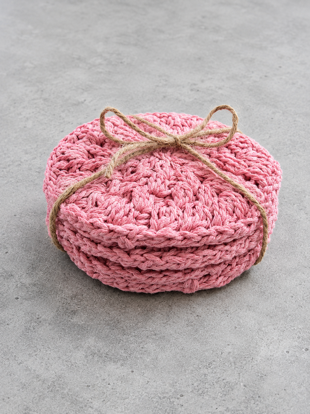 A stack of round pink crocheted dishcloths tied with a natural jute twine bow, set against a textured gray concrete background.