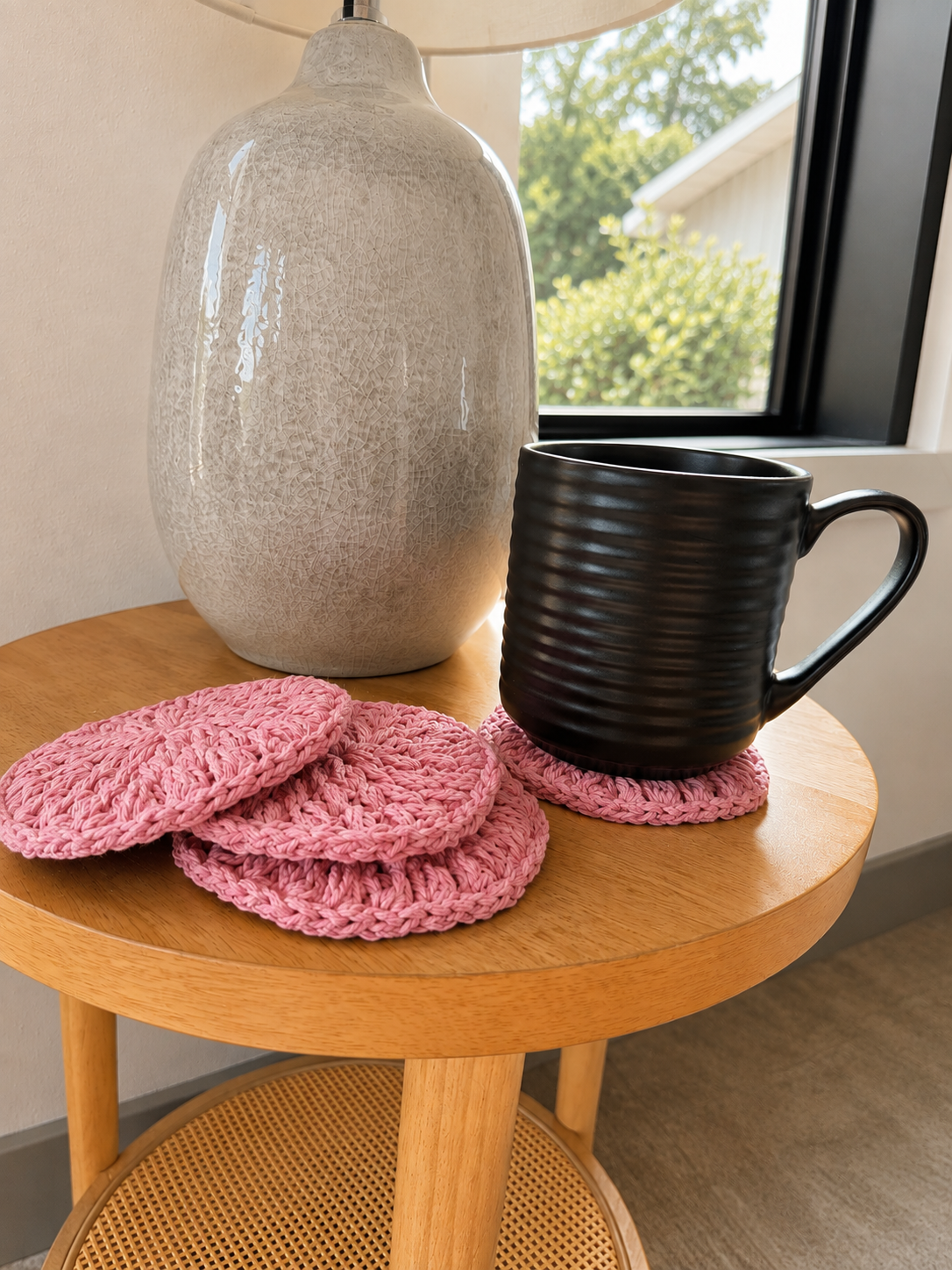 A set of pink crocheted coasters on a round wooden side table, styled with a black ribbed ceramic mug and a large textured gray vase in front of a window.