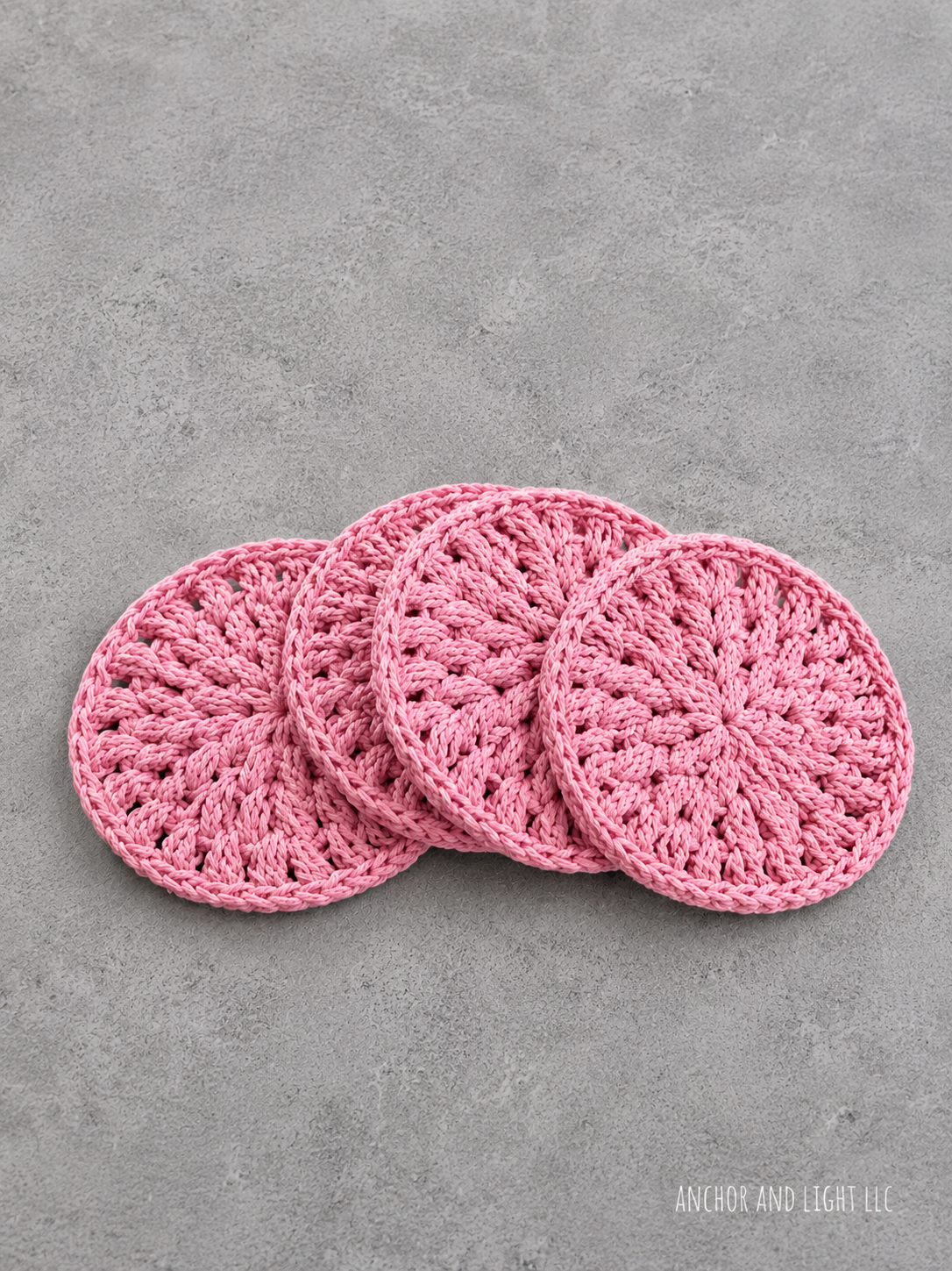 Four pink crocheted round coasters arranged in an overlapping fan shape on a textured gray concrete surface. The brand name 'ANCHOR AND LIGHT LLC' is visible in the bottom right corner.