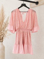 A pink lace and satin babydoll dress with wide flutter sleeves and a smocked waist, hanging on a black hanger against a textured beige wall with dried grass decor.