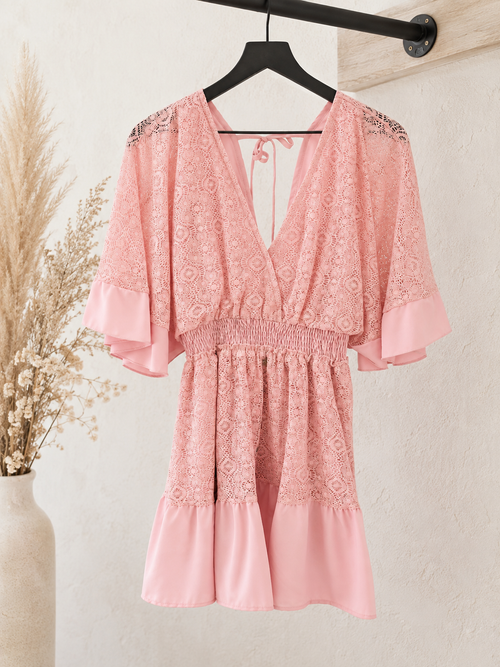 A pink lace and satin babydoll dress with wide flutter sleeves and a smocked waist, hanging on a black hanger against a textured beige wall with dried grass decor.