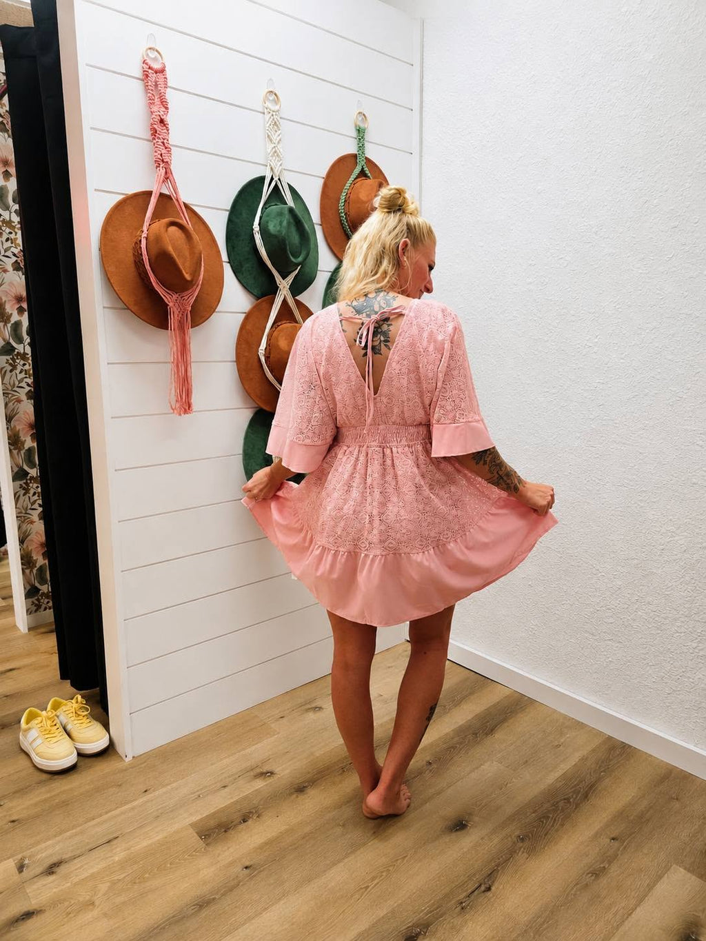 A woman stands with her back to the camera wearing a light pink lace mini dress with elbow-length sleeves and a ruffled hem, in a room with wood floors and white shiplap walls with hanging hats.