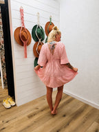 A woman stands with her back to the camera wearing a light pink lace mini dress with elbow-length sleeves and a ruffled hem, in a room with wood floors and white shiplap walls with hanging hats.