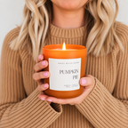 A person in a beige knit sweater holds a lit orange candle jar labeled 'Pumpkin Pie' by Sweet Water Scents against a soft light gray background.