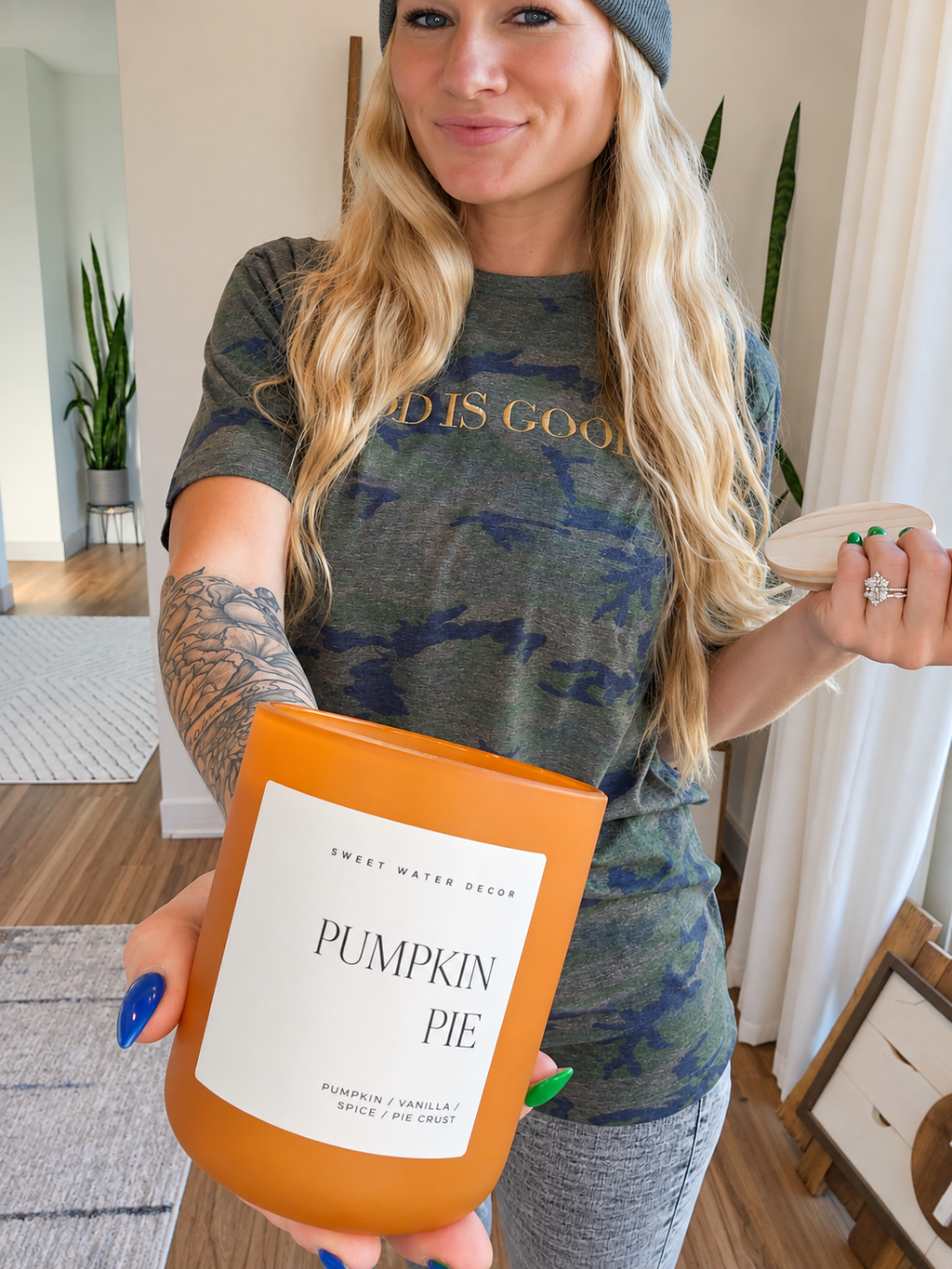 A woman holding a 'Pumpkin Pie' scented candle in an orange glass jar, wearing a camouflage t-shirt and beanie in a bright indoor setting.