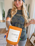 A woman holding a 'Pumpkin Pie' scented candle in an orange glass jar, wearing a camouflage t-shirt and beanie in a bright indoor setting.