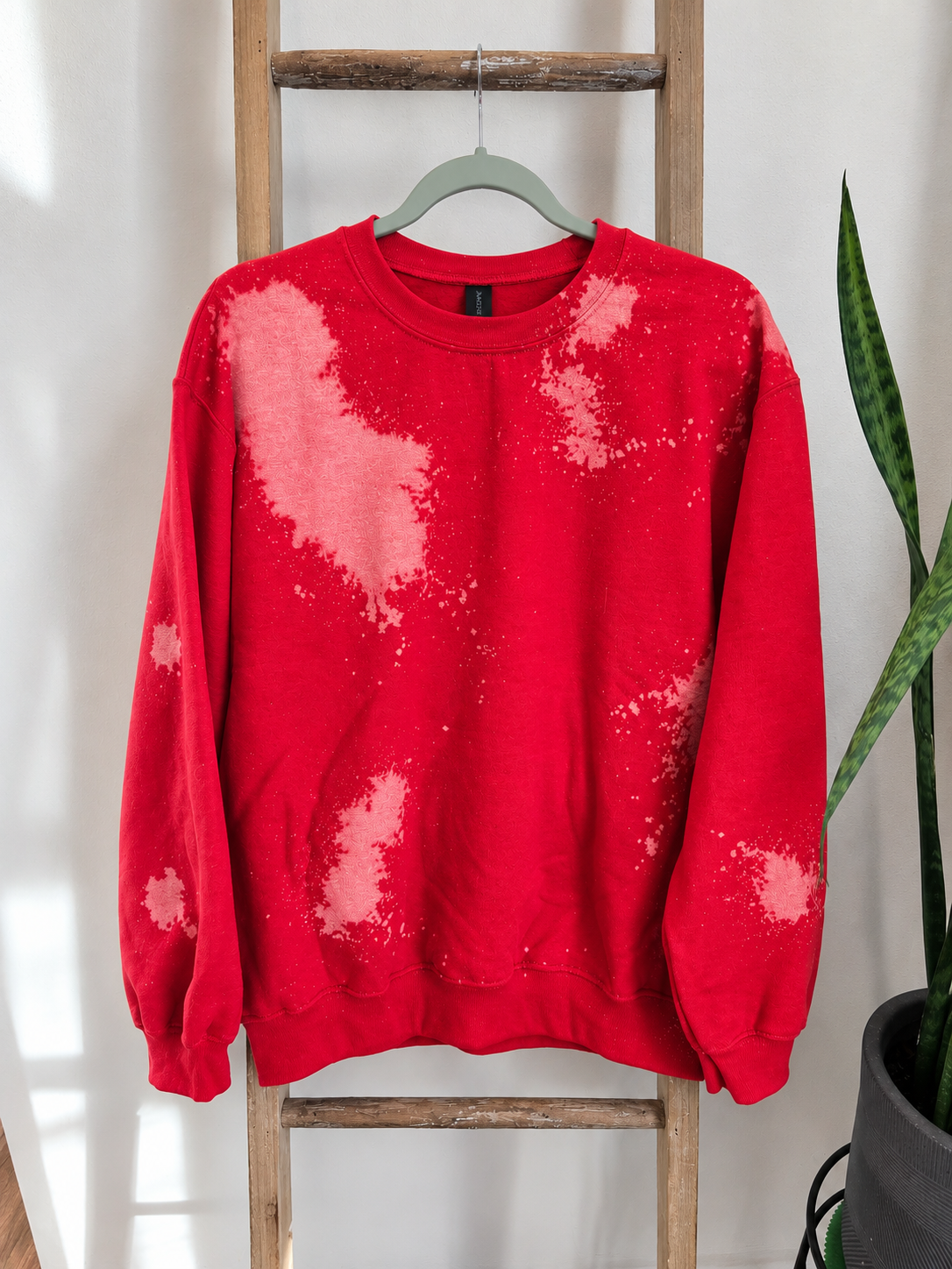 A red crewneck sweatshirt with a pink and white bleach-dye splatter pattern, hanging on a light green hanger on a rustic wooden ladder next to a potted snake plant.