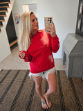 A woman takes a mirror selfie wearing a bright red crewneck sweatshirt with white bleach-splatter details and white distressed denim shorts in a home setting.