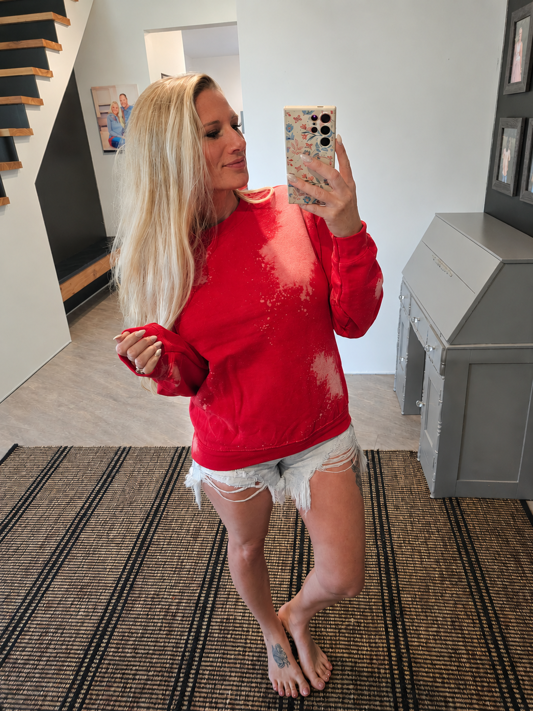 A woman takes a mirror selfie wearing a bright red crewneck sweatshirt with white bleach-splatter details and white distressed denim shorts in a home setting.