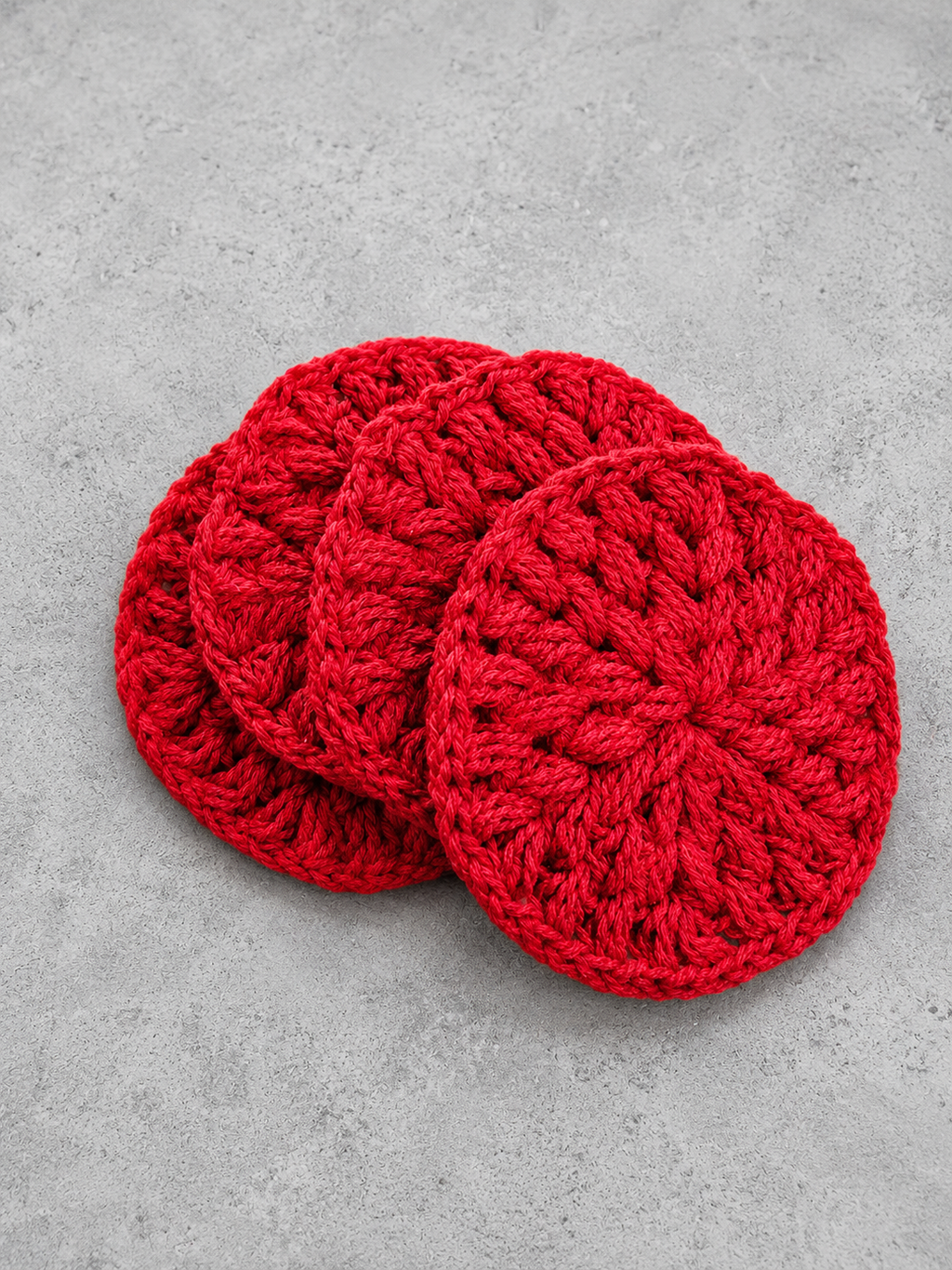 Four round red crocheted coasters stacked slightly offset on a textured gray surface.