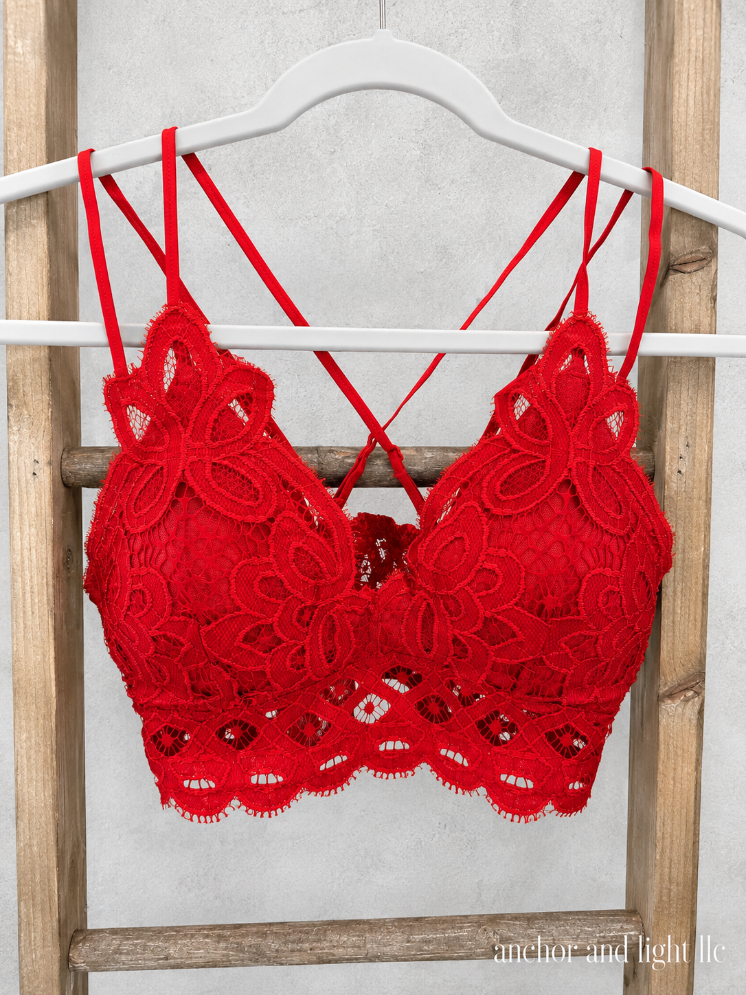 A bright red lace bralette with floral lace detailing and thin crossover back straps, hanging on a white hanger against a rustic wooden ladder backdrop.