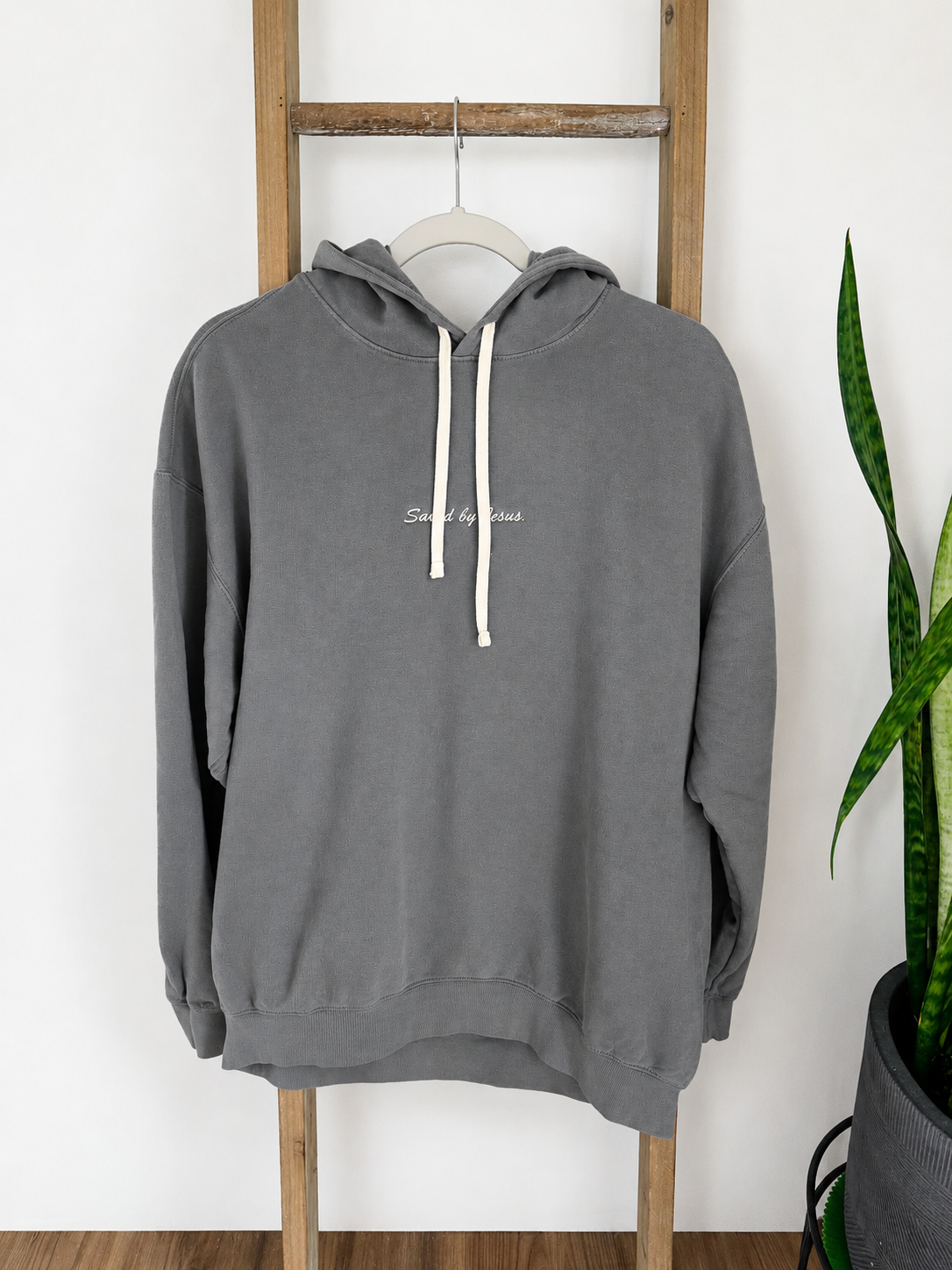 A dark gray hooded sweatshirt with cream drawstrings and small white script text on the chest, hanging on a wooden ladder against a white wall with a green plant visible on the right.