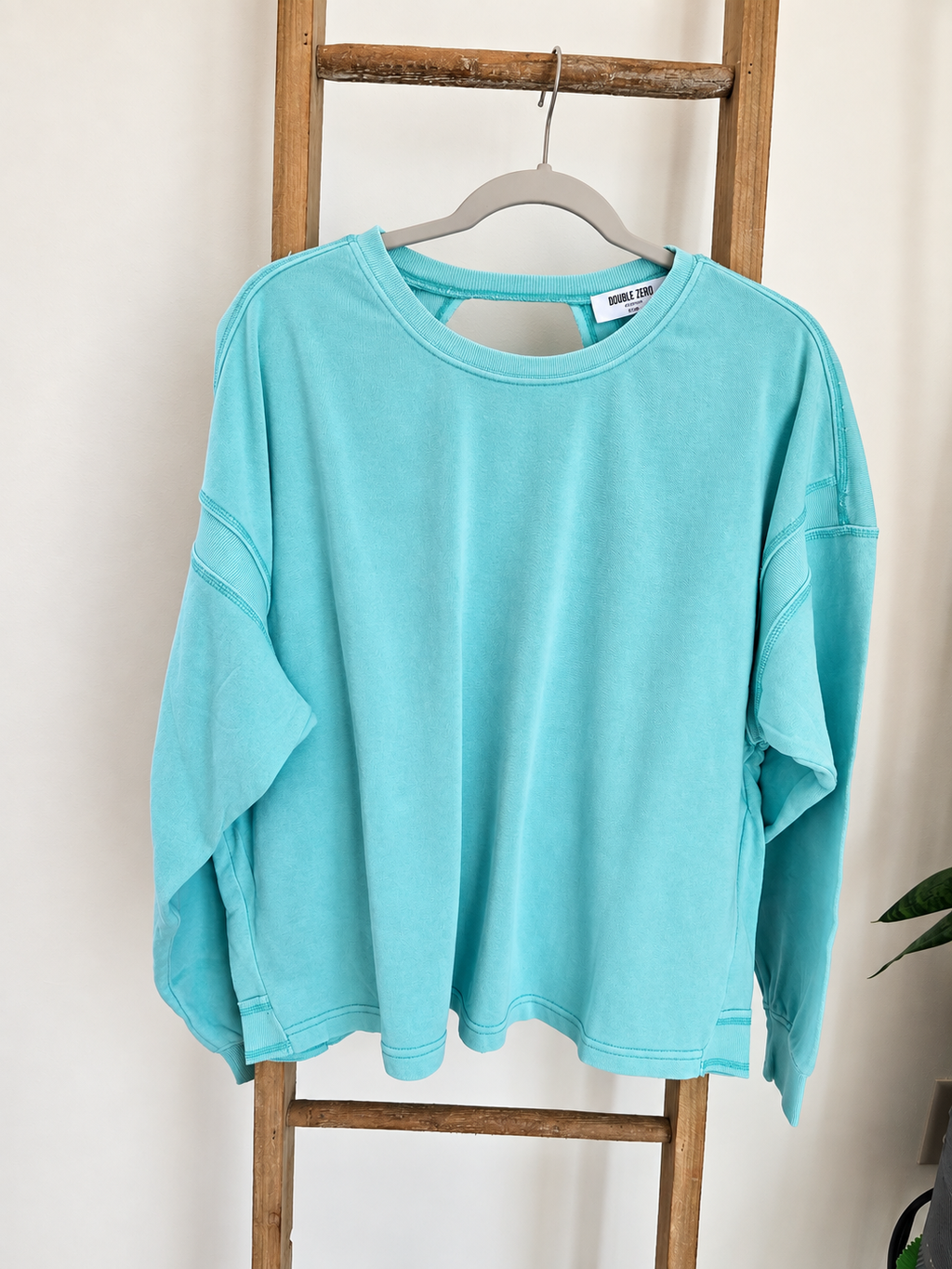 A turquoise long-sleeve cropped sweatshirt with a keyhole cutout at the neck, hanging on a gray hanger from a rustic wooden ladder against a white wall.