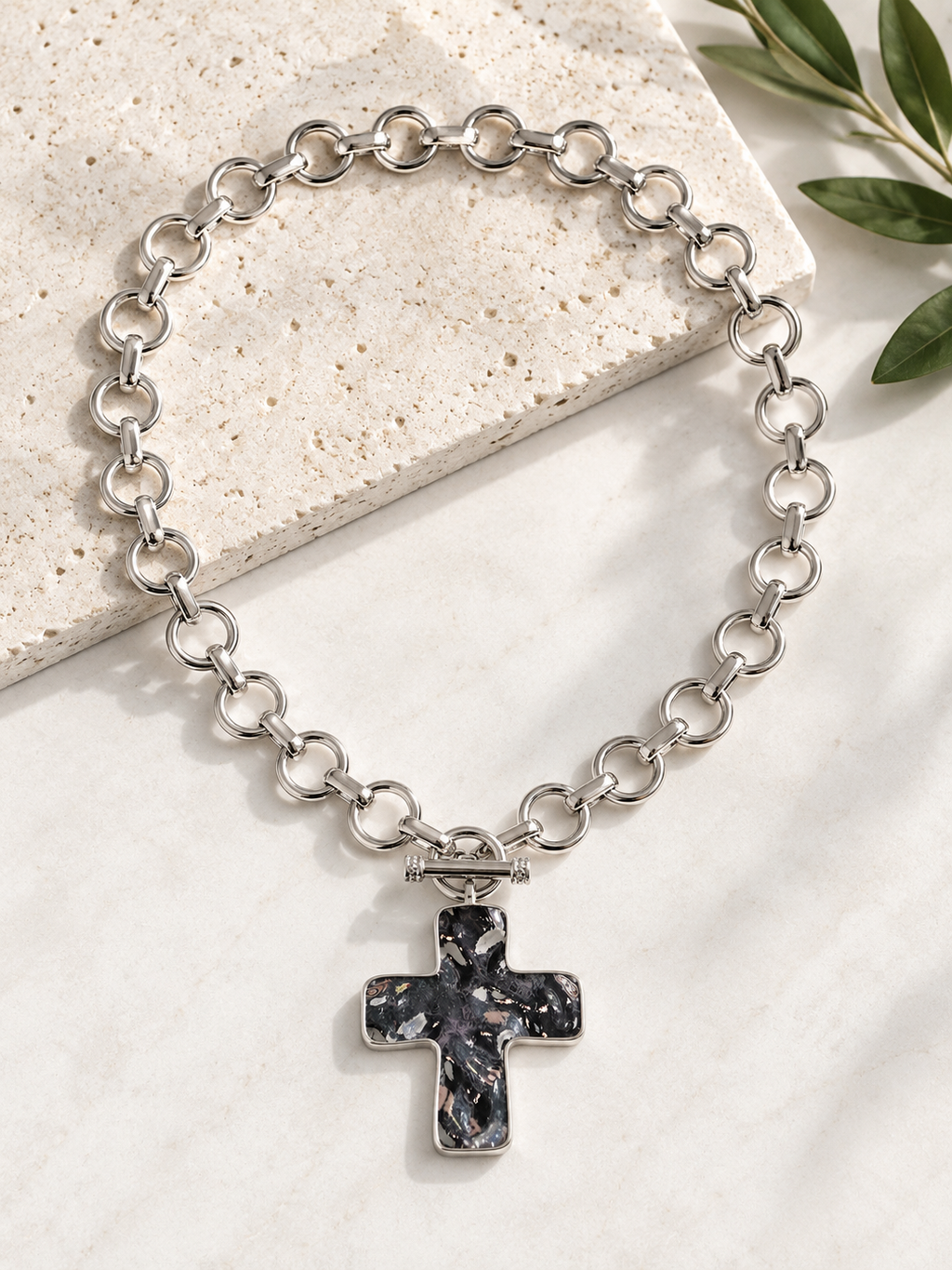 A silver chain bracelet with a cross pendant featuring a black and white marbled stone inlay, styled on a light stone slab with green leaves in the corner.
