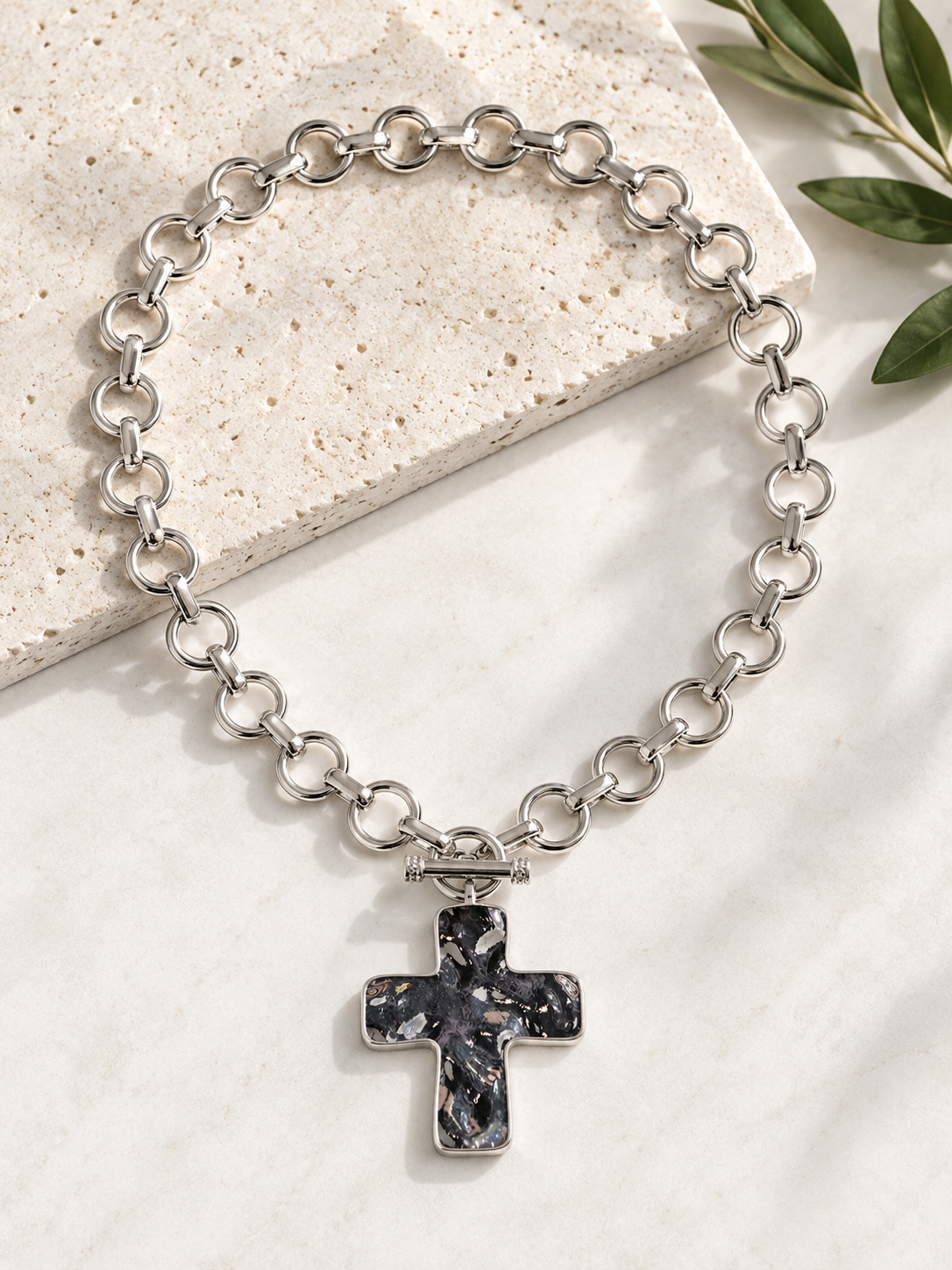 A silver chain bracelet with a cross pendant featuring a black and white marbled stone inlay, styled on a light stone slab with green leaves in the corner.