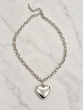 A silver chain necklace with a polished heart-shaped pendant, displayed on a white marble surface.