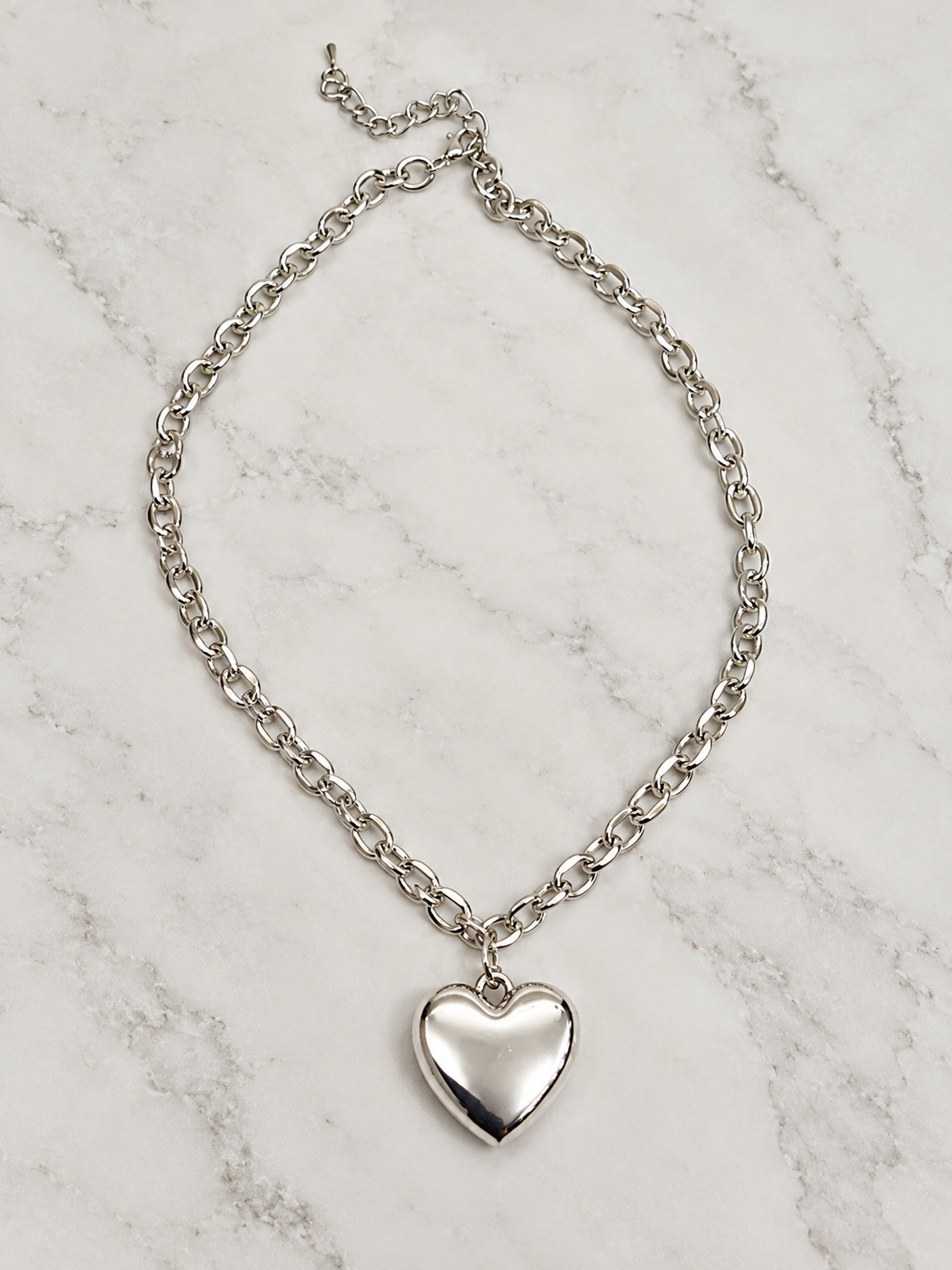 A silver chain necklace with a polished heart-shaped pendant, displayed on a white marble surface.