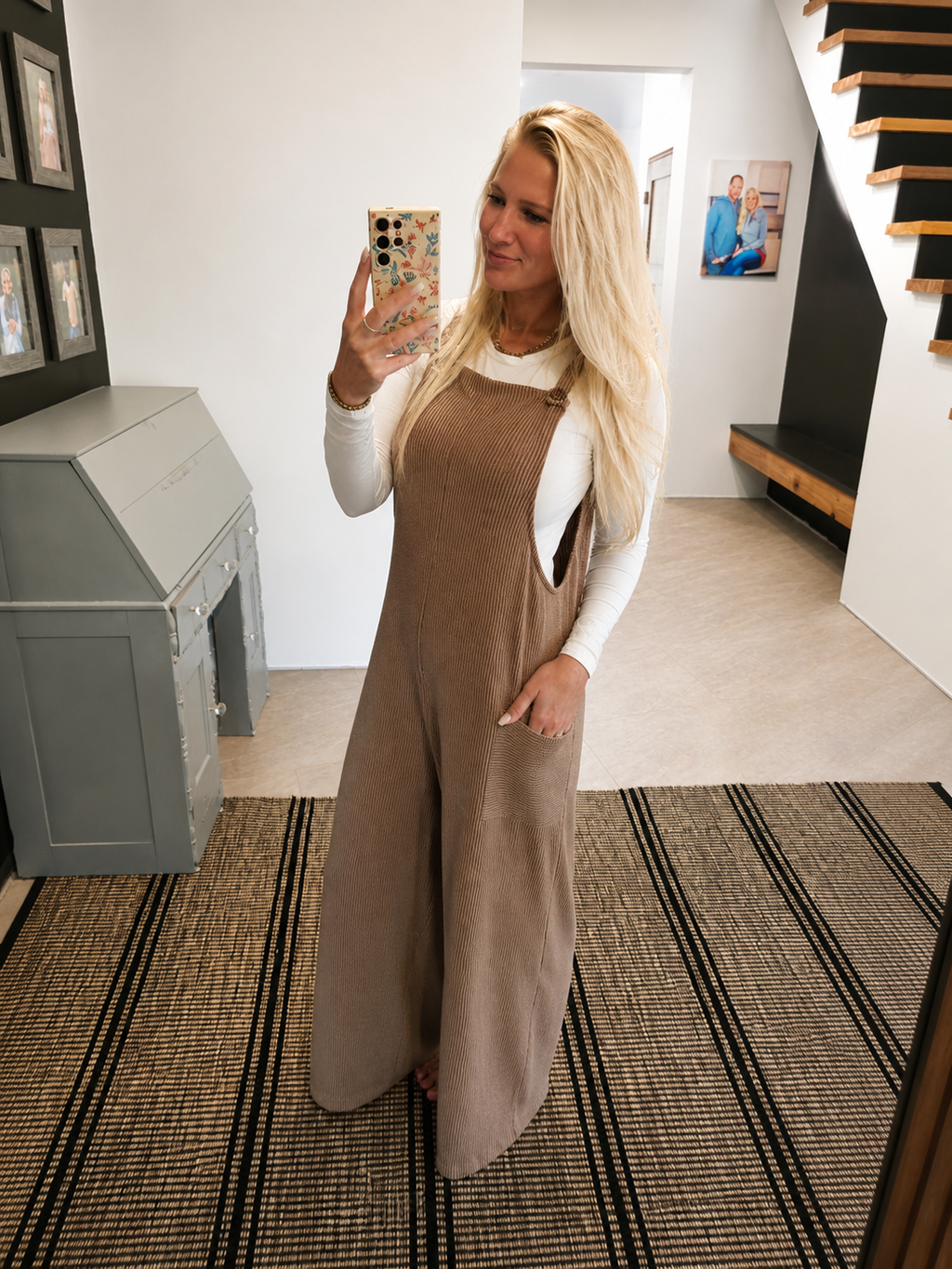 A woman takes a mirror selfie in a home setting, wearing a brown corduroy jumpsuit over a white long-sleeve top. The jumpsuit features a bib front, side pockets, and a relaxed, wide-leg fit.