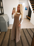 A woman takes a mirror selfie in a home setting, wearing a brown corduroy jumpsuit over a white long-sleeve top. The jumpsuit features a bib front, side pockets, and a relaxed, wide-leg fit.
