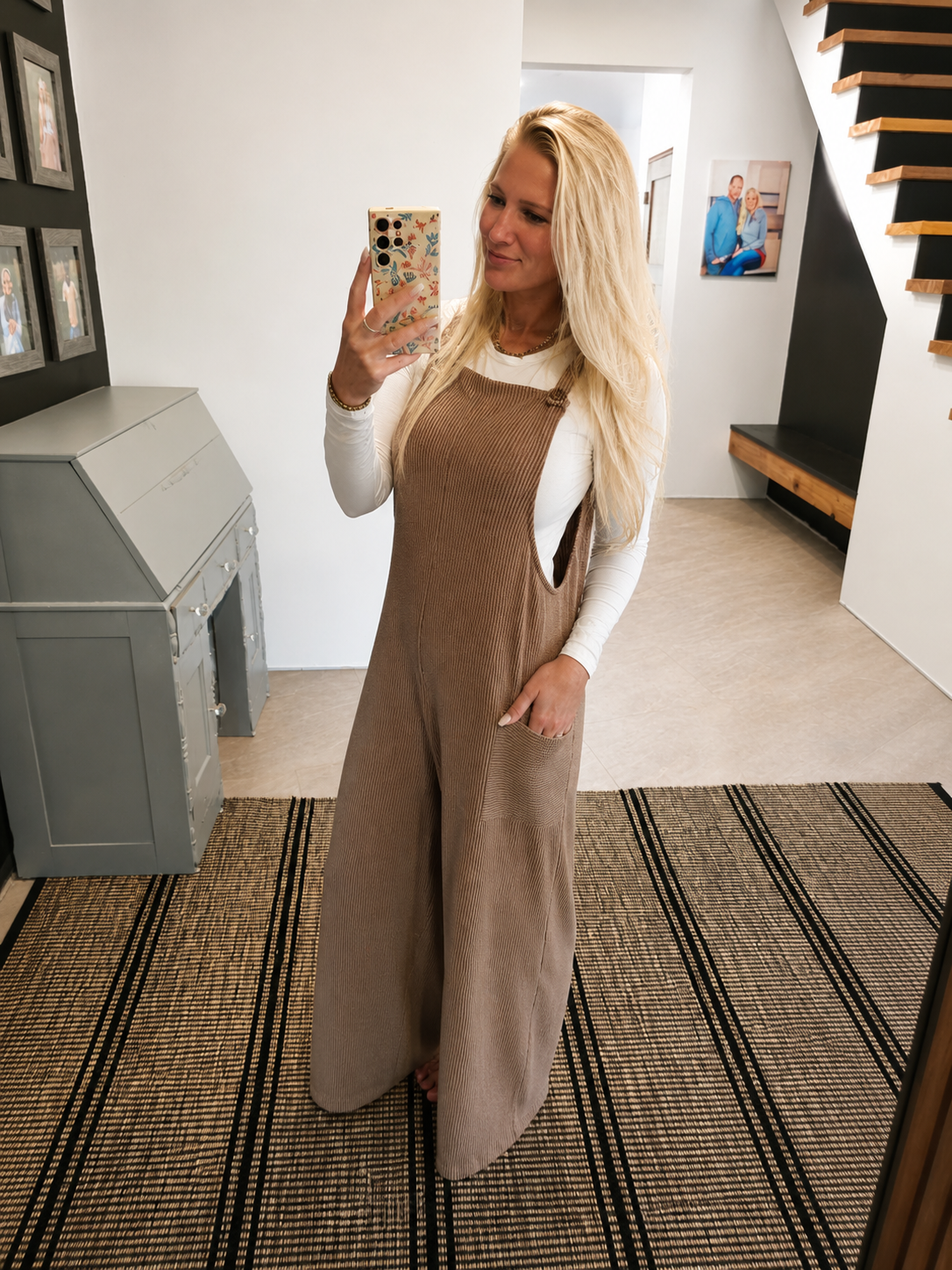 A woman takes a mirror selfie in a home setting, wearing a brown corduroy jumpsuit over a white long-sleeve top. The jumpsuit features a bib front, side pockets, and a relaxed, wide-leg fit.