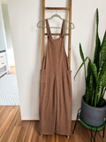 A pair of brown corduroy bib overalls hanging on a wooden ladder in a home setting, with a snake plant in a gray pot on a green stand to the right.