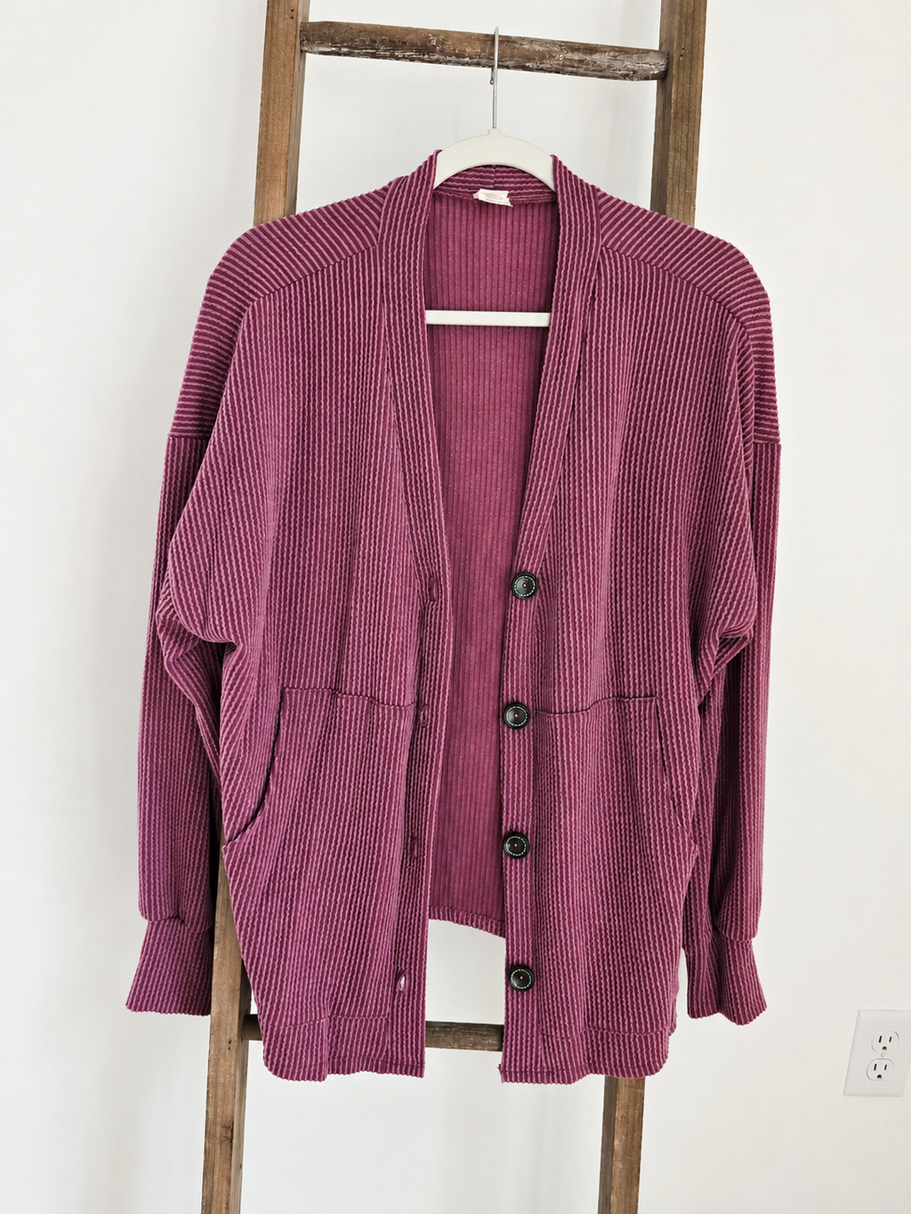 A ribbed knit cardigan sweater in a dusty mauve-purple color, hanging on a white hanger on a wooden ladder against a white wall.