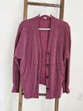 A ribbed knit cardigan sweater in a dusty mauve-purple color, hanging on a white hanger on a wooden ladder against a white wall.