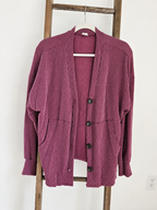 A ribbed knit cardigan sweater in a dusty mauve-purple color, hanging on a white hanger on a wooden ladder against a white wall.