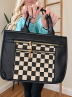 A person holds up a black faux-leather tote bag with a black and cream checkered front pocket and gold-tone zippers. The person is wearing a light blue sweatshirt and dark pants, standing in an indoor room with a wooden ladder and plant in the background.