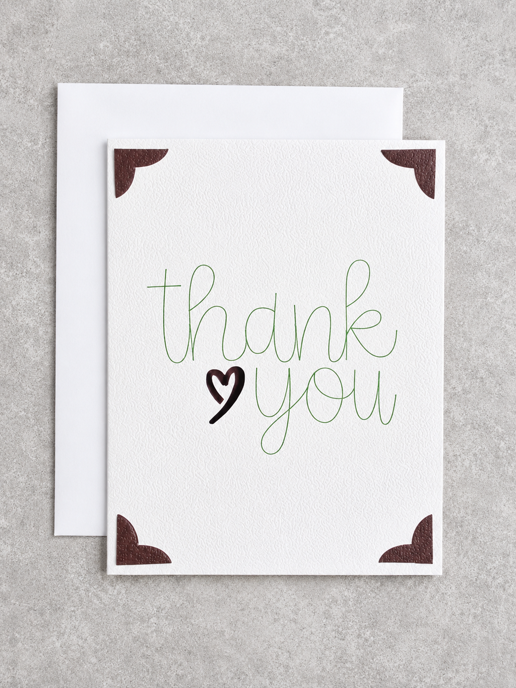 A square white greeting card with the words 'thank you' in green script and a black heart, shown with a white envelope behind it on a light gray textured background.