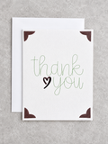 A square white greeting card with the words 'thank you' in green script and a black heart, shown with a white envelope behind it on a light gray textured background.