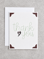 A square white greeting card with the words 'thank you' in green script and a black heart, shown with a white envelope behind it on a light gray textured background.
