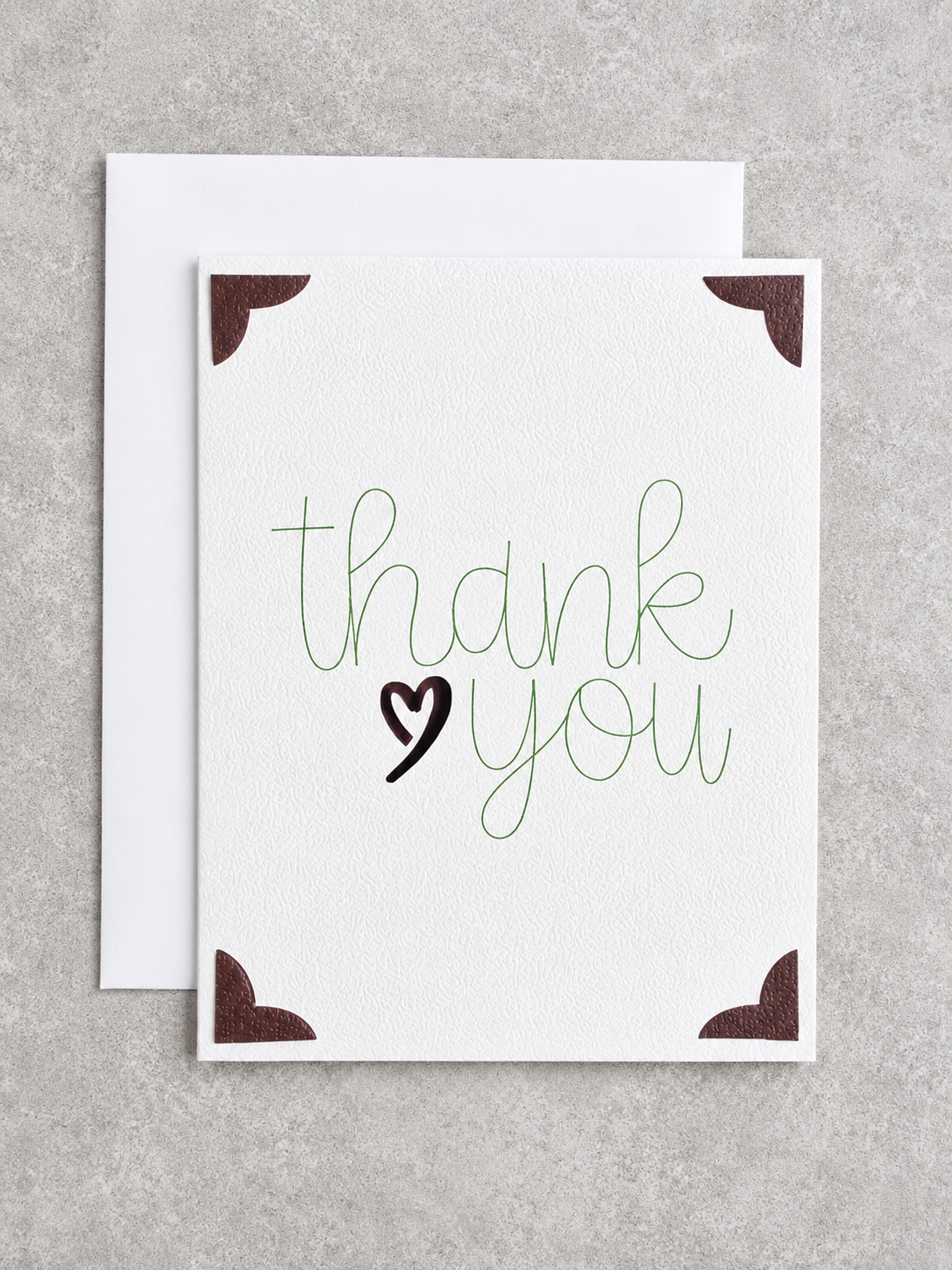 A square white greeting card with the words 'thank you' in green script and a black heart, shown with a white envelope behind it on a light gray textured background.