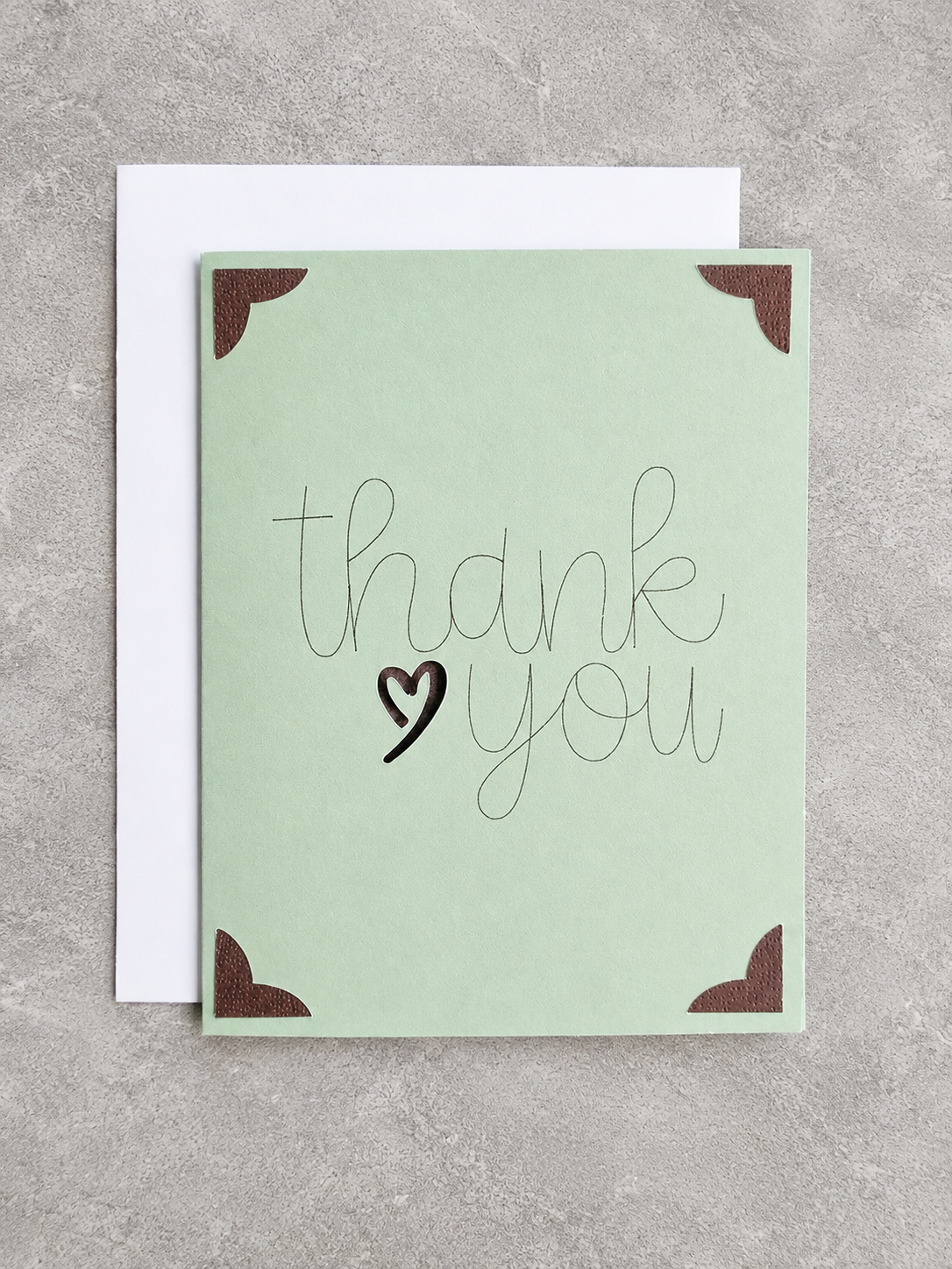 A square thank you card in pale mint green with brown corner accents and the words 'thank you' in script, shown with a white envelope on a gray textured background.