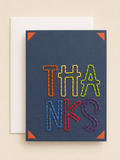 A square navy blue greeting card with the word 'THANKS' spelled out in colorful, thread-like letters, shown with a white envelope behind it on a warm beige background.